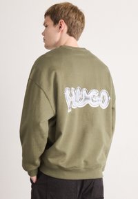 Młody mężczyzna stoi bokiem, ubrany w oliwkową bluzę z połyskującym białym napisem "HUGO" na plecach i ciemne spodnie, trzymając ręce w kieszeniach.