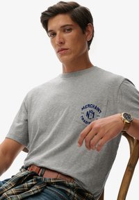 Camiseta de algodón gris con mangas cortas, con un estampado gráfico azul en el bolsillo izquierdo del pecho y un corte casual. Lleva un cinturón con una camisa a cuadros atada a la cintura.