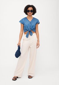 Camisa de ganga cropped de mangas curtas com detalhe em nó na cintura, combinada com calças largas em creme e uma bolsa texturizada em azul-marinho.