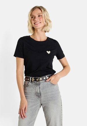 T-shirt en coton noir à manches courtes, encolure ronde et petit détail en forme de cœur doré sur la poitrine, associé à un jean gris clair et une ceinture noire.