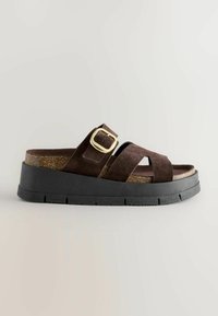 Sandelă din suede maro cu o talpă neagră masivă, un miez din plută și un accent din aur pe curea. Dispune de curele încrucișate pentru o fixare sigură.