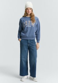 Felpa azzurra con la scritta "CREW" in una fantasia leopardata, abbinata a jeans a gamba larga blu e sneakers bianche; completata da un berretto di maglia color crema.