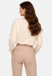 Femme aux longs cheveux bruns ondulés portant un pull crème et un pantalon beige taille haute, vue de dos sur un fond uni.