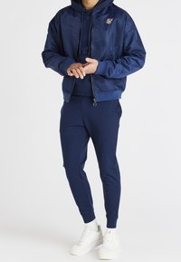 Marinblå bomberjacka med huva, dragkedja och logotyp; matchas med smala sweatpants i samma färg. Vita sneakers kompletterar outfiten.