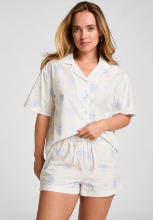 Vrouw draagt een kort mouw knoopblouse en bijpassende korte broek met lichtblauw bloemenpatroon, staand tegen een effen achtergrond.