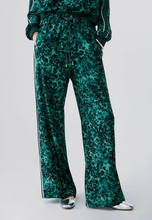 Persona que lleva pantalones verdes sueltos con patrón, cintura elástica y rayas laterales blancas, combinados con zapatos plateados apuntados.