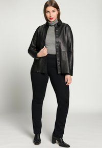 Schwarze Lederjacke mit Kragen-Design, Knopfverschluss und langen Ärmeln, getragen über einem grauen Rollkragenpullover und schwarzen, eng anliegenden Hosen.