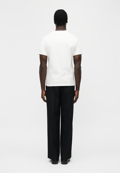 Homme debout, de dos, portant une chemise blanche unie à manches courtes, un pantalon noir à jambes larges et des chaussures noires sur un fond blanc uni.