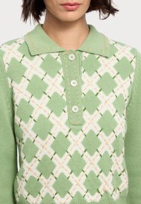 Grøn og hvid strikket polo, med diamantmønster og orange accenter. Fire knapper og kravehals, ribbet tekstur på ærmerne.