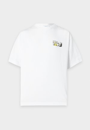 Homme mince à la peau foncée portant un t-shirt graphique blanc oversize, un short cargo gris clair, des baskets noires, des chaussettes blanches et des lunettes de soleil noires, debout.