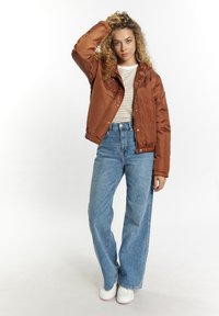 Brun, polstret jakke med lynlås og trykknaplukning, parret med højhalsede lysblå jeans og hvide sneakers, sat mod en ensfarvet baggrund.