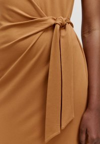 Détail d'une robe beige avec un tissu doux, un accent noué à la taille, une texture lisse et une silhouette fluide.