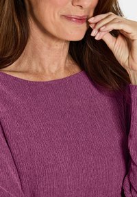 Frau mit langen braunen Haaren, die ein strukturiertes lila Langarmshirt trägt und mit ihrer rechten Hand ihr Gesicht in der Nähe der Lippen berührt.