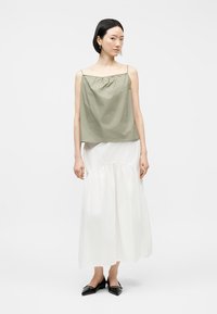 Γυναίκα όρθια, φοράει φαρδιά χακί αμάνικη μπλούζα, μακριά λευκή φούστα και μαύρες μυτερές μπαλαρίνες μπροστά σε απλό λευκό φόντο.