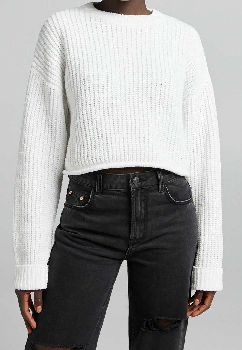 Pull en maille blanc avec un col rond et une texture côtelée, associé à un jean noir taille haute avec des détails déchirés.