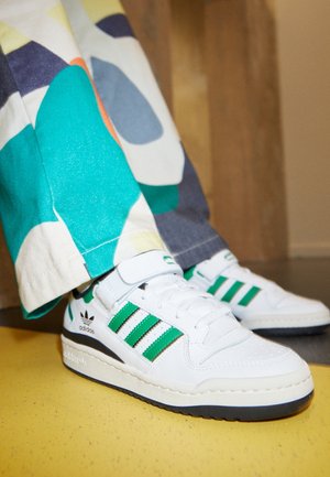 Białe sneakersy Adidas z zielonymi paskami noszone z wielokolorowymi spodniami, stojące na żółtej podłodze obok drewnianej powierzchni.