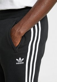 Svarta träningbyxor med vitt tre-stripesmönster längs sidorna, med en broderad logotyp och en framficka. Tillverkade av mjukt tyg.