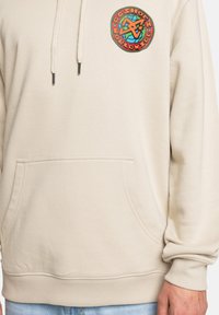 Béžová mikina s predným vreckom, kapucňou na šnúrku a farebnou kruhovou grafikou s globusom a textom. Jemná, štruktúrovaná tkanina.