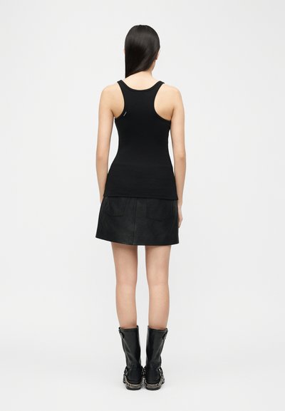 Femme aux longs cheveux foncés portant un débardeur noir ajusté à dos nageur, une jupe en cuir noir et des bottes noires mi-mollet, debout de dos.