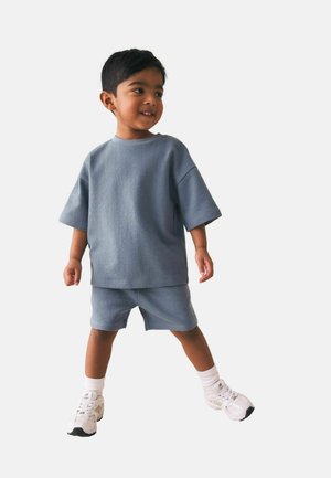 Noor poiss seisab jalad harkis, seljas lõdvalt istuv sinine lühikeste varrukatega särk ja sobivad püksid, valged sokid ja valged tossud.