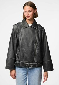 Veste en similicuir noir avec un grand col, une taille ceinturée et des manches larges. Des coutures contrastées et des poches latérales sont visibles.