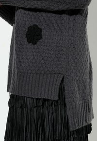Pull en maille grise avec un motif texturé et un ourlet côtelé, orné d'une broderie florale noire sur le côté, associé à une jupe noire à franges.