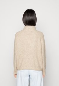 Beige gestrickter Pullover mit hohem Kragen, lockerer Passform und strukturierter Oberfläche, kombiniert mit hellblauen Hosen, von hinten gezeigt.
