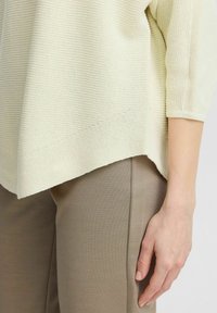 Helles gelbes Strickoberteil mit strukturiertem Muster, mit drei Viertelärmeln und einem gespaltenen Saum, kombiniert mit beigen, figurbetonten Hosen.