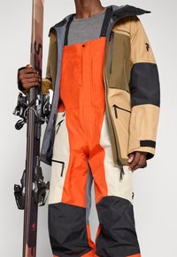 Suusariietus sisaldab multivärvilist veekindlat jakki beeži, halli ja musta värviga, millele on sobitatud ereoranž ja kreemjas kombinesoon ning suusapulk.