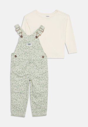 Chemise crème à manches longues associée à une salopette verte à motifs floraux avec bretelles réglables à boucle et boutons latéraux.