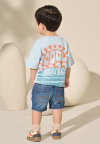 Mlad otrok stoji obrnjeno stran, oblečen v svetlo modro majico z oranžnim in modrim napisom "Cool Sunny Days" ter jeans kratke hlače v nevtralnem okolju.