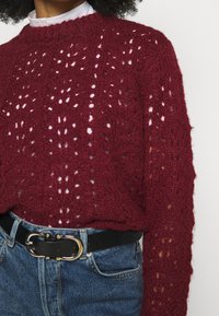 Maglione lavorato a maglia bordeaux con un motivo aperto testurizzato, abbinato a una cintura nera con una fibbia a doppio anello dorato e jeans di denim.