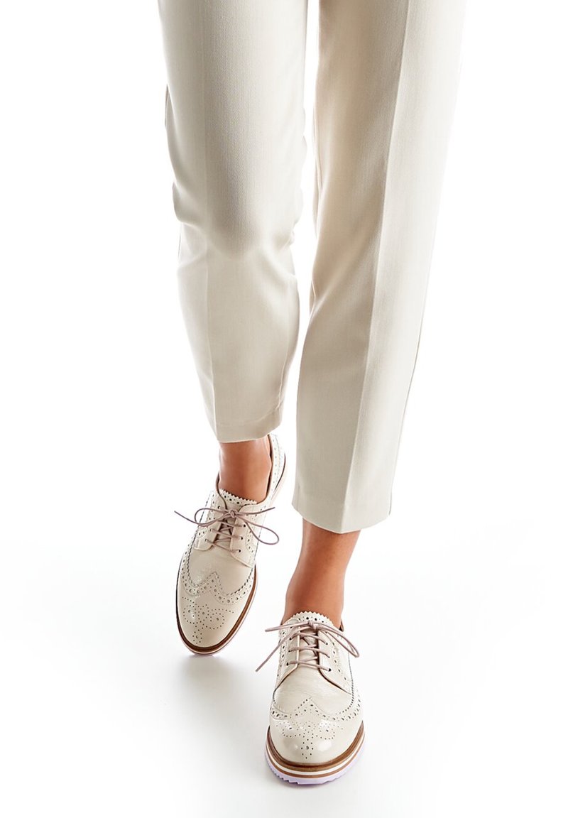 Brogues en cuir beige avec des détails perforés, des lacets crème et une semelle blanche, associés à un pantalon tailleur de couleur claire.