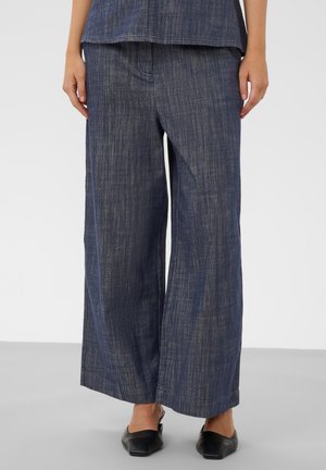 Femme portant un pantalon large bleu ample et des chaussures noires à bout pointu sans lacets, debout sur un sol gris.