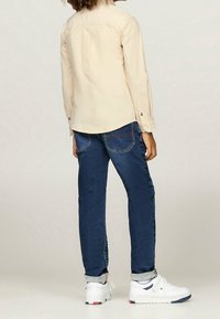 Osoba w beżowej koszuli z długim rękawem, ciemnoniebieskich dżinsach z mankietami i białych trampkach, stojąca tyłem na jednolitym tle.