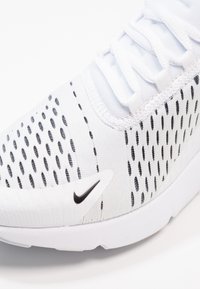 Weißer Sportschuh mit einem atmungsaktiven Mesh-Obermaterial, das mit schwarzen Punktmustern, einem schwarzen Logodetail und einer strukturierten Sohle versehen ist.