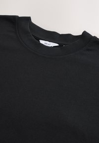 Sweat-shirt noir à col rond avec étiquette de marque visible à l'intérieur du col sur un fond clair.