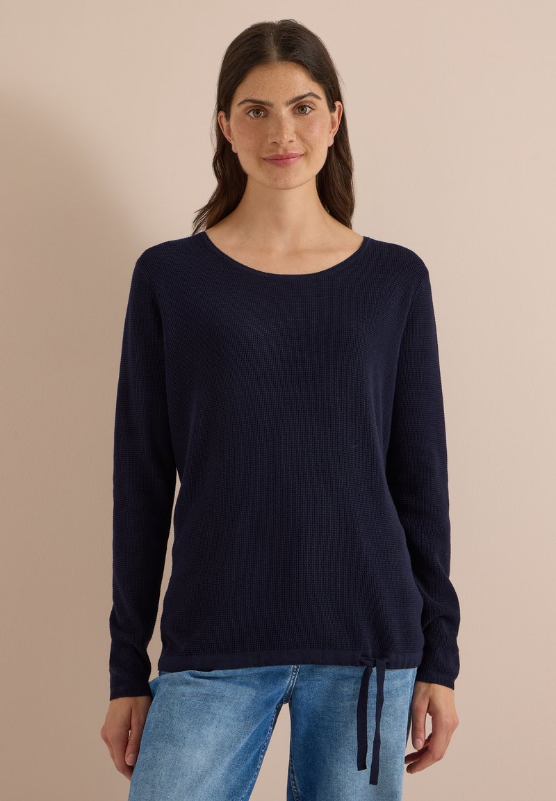 Femme aux cheveux châtain foncé portant un pull bleu marine à manches longues et un jean bleu clair, debout devant un fond beige uni.