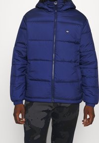 Marinblå pufferjacka med dragkedja fram, horisontell sömnad och huva. Har en liten logotyp på bröstet och ribbade ärmslut.