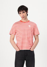 Tricou cu mânecă scurtă în dungi roșii și albe, cu guler rotund și un emblema albă cu floare pe partea stângă a pieptului. Purtați-l cu blugi negri.