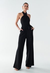 Combinaison noire à col halter en tissu texturé, avec une coupe évasée, des poches latérales et une ceinture à nouer à la taille. Accentuée par des boucles d'oreilles circulaires dorées.