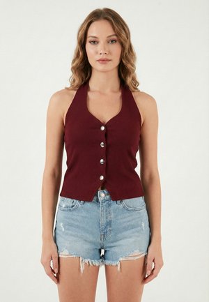 Kobieta ubrana w bordowy, bezrękawnik na guziki z wiązaniem na szyi i pastelowo niebieskie postrzępione szorty jeansowe, stojąca na jednolitym tle.