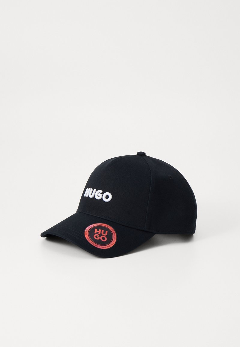 Svart baseballcap laget av stoff, med hvit brodert "HUGO" logo foran og en rød sirkulær detalj med "HUGO" på siden.