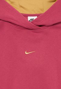 Mikina Nike v hlbokej ružovej farbe so zlatou vyšitou logom swoosh. Obsahuje vrecko typu kangaroo a mäkkú vnútornú podšívku v svetlejšom odtieni.
