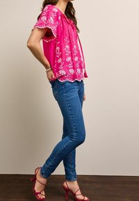 Rosa, kurzärmliges Oberteil mit weißer Blumenstickerei, kombiniert mit blauen enganliegenden Jeans und rosa Riemchen-Pumps. Flacher, strukturierter Holzboden.