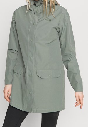 Woman wearing a light green hooded rain jacket with snap buttons and front flap pockets, paired with black leggings.