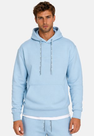 Homme portant un sweat à capuche bleu clair et un pantalon de survêtement assorti avec cordons, debout devant un fond uni.