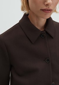 Bruin jack met knopen en een gestructureerde kraag, voorzien van zwarte knopen, een gladde textuur en een getailleerde pasvorm. Close-up van het halsgebied.