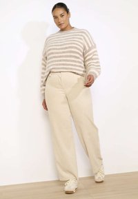 Bež prugast pleten pulover z sproščenim krojem in spuščeni rameni, v kombinaciji z svetlobezž širokimi hlačami iz kordirane tkanine in kremnimi športnimi čevlji.