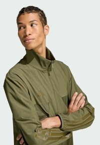 Olive green lightweight jacket with a high collar, zip closure, and two front pockets. Features stripes on sleeves and subtle branding.